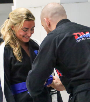 Smiling woman in a black gi with a white belt shaking hands with a bald man in a black gi during a martial arts class.