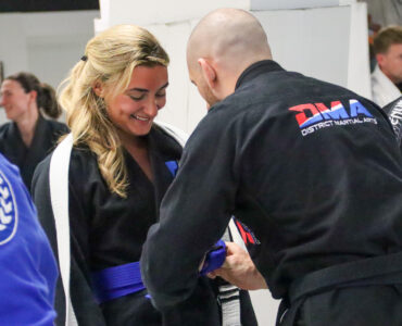 Smiling woman in a black gi with a white belt shaking hands with a bald man in a black gi during a martial arts class.