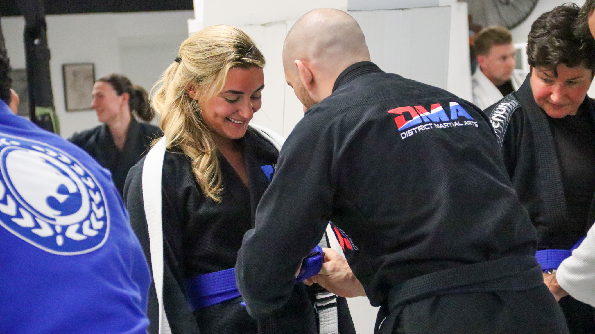 Smiling woman in a black gi with a white belt shaking hands with a bald man in a black gi during a martial arts class.