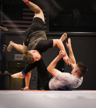 Male fighter in black shorts leaps overhead inside a cage as teammates reach up to assist; an American flag visible in the background.