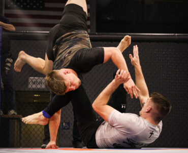 Male fighter in black shorts leaps overhead inside a cage as teammates reach up to assist; an American flag visible in the background.