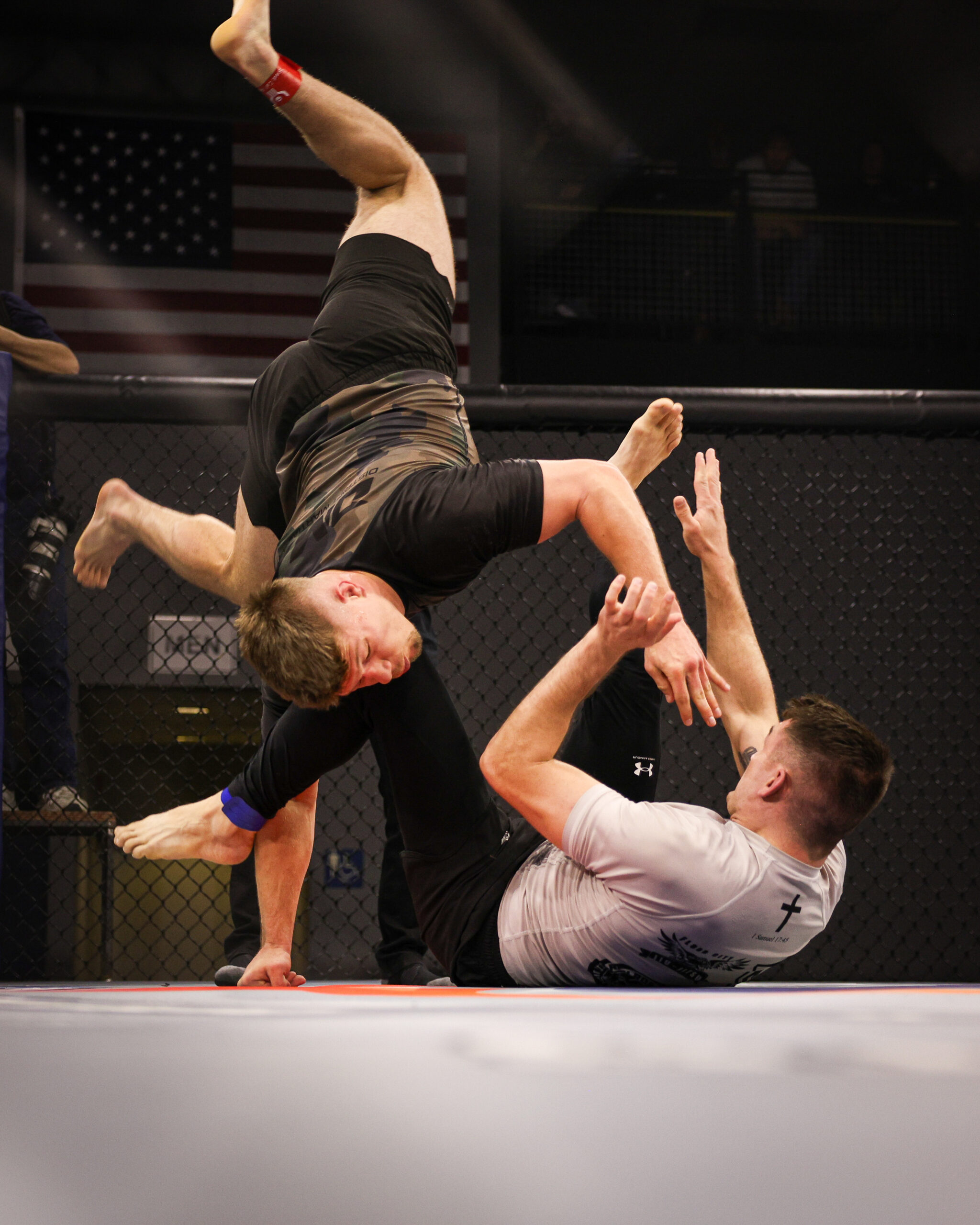 Male fighter in black shorts leaps overhead inside a cage as teammates reach up to assist; an American flag visible in the background.