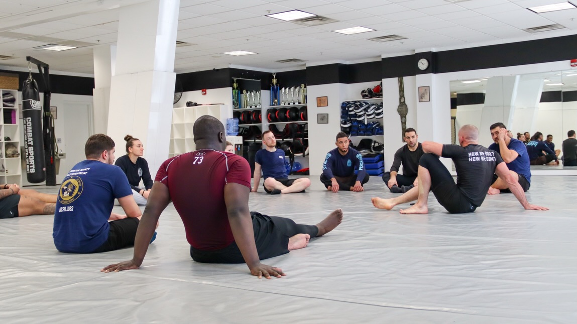 Group of people in a martial arts training gym sitting in a circle on gray mats, listening to instruction.