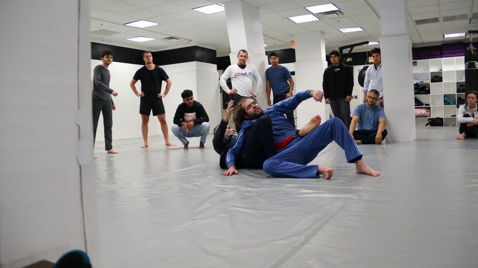 Two people grapple on a padded gym mat while a group of students watches from around the edge of the mat.