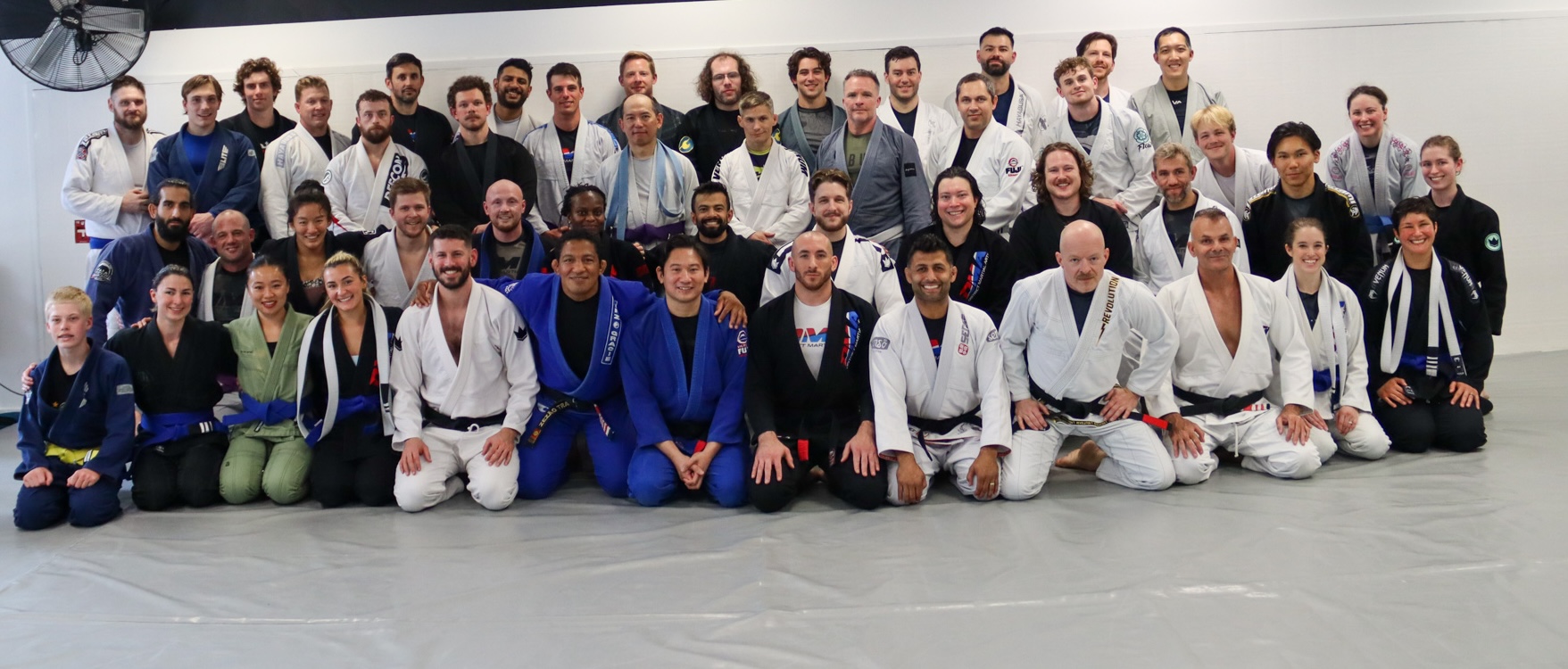 Group of men and women in BJJ gis posing for a class photo on a gray mat in a dojo