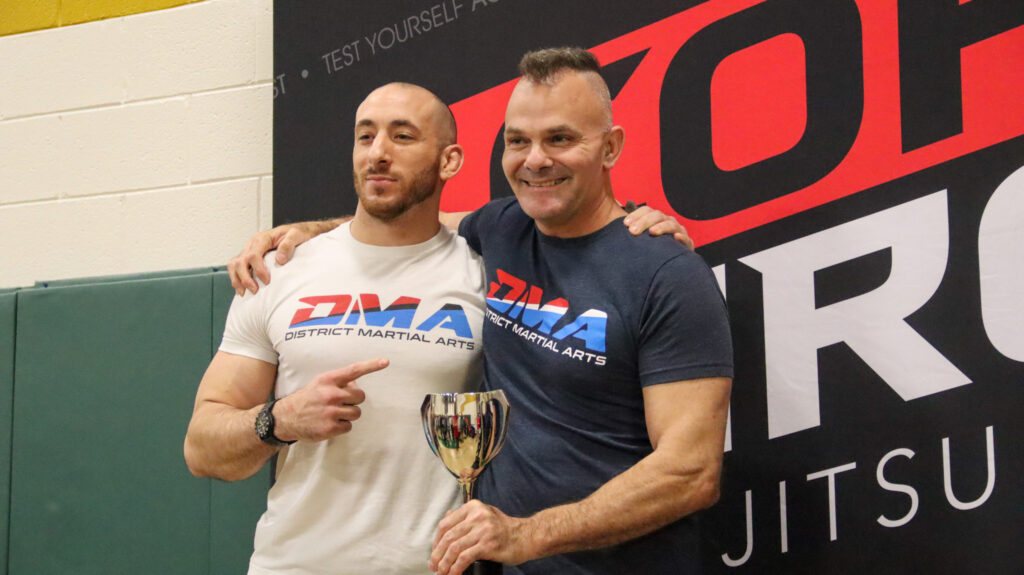 Two men posing together after a martial arts event, one in a white DM A shirt and the other in a navy DM A shirt, holding a trophy with a large banner behind them G