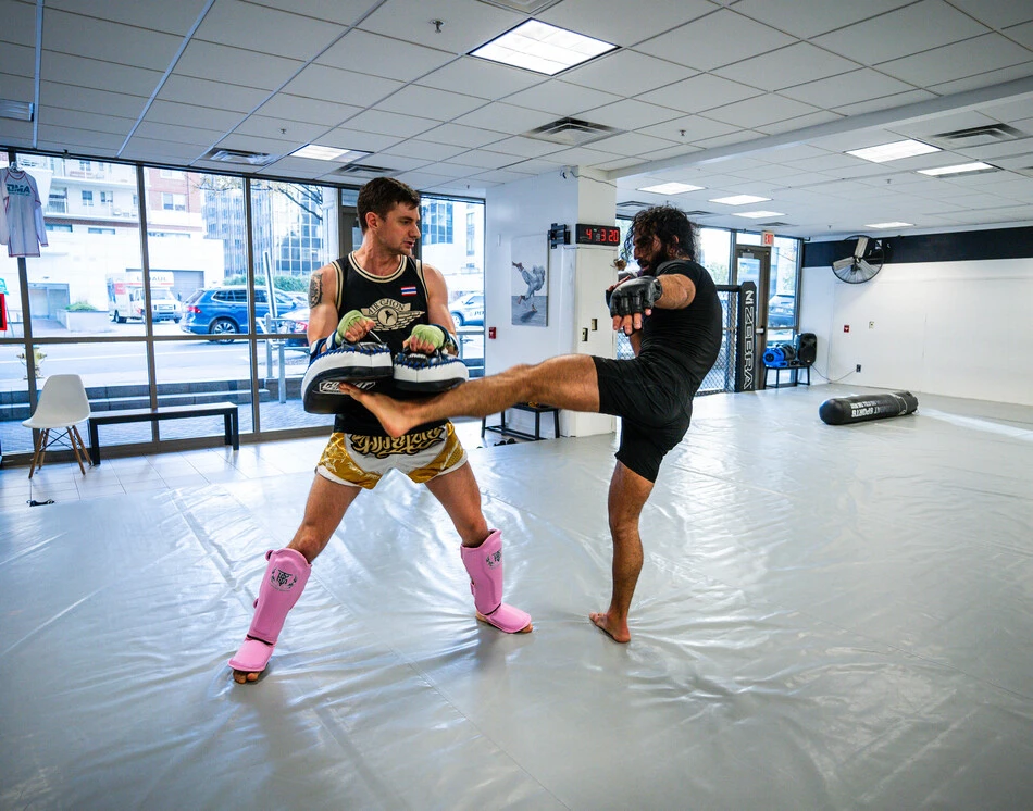 Two mixed martial artists spar in a bright gym; one holds pink shin guards and pads while the other delivers a high kick toward the pad holder.