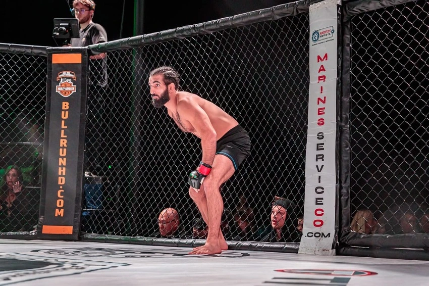 MMA fighter with red gloves crouches in a fenced fighting cage, ready to fight.