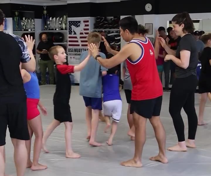 Group of kids and adults in a gym high-fiving during a Muay Thai class; banner reading USA Muay Thai and trophies visible in the background.